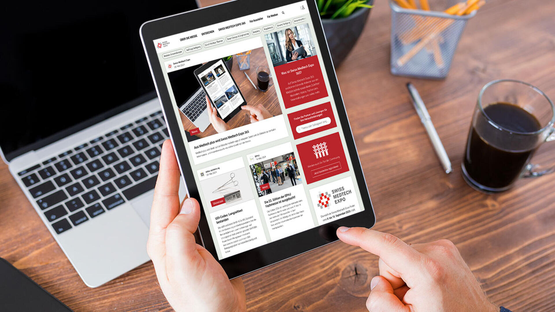Eine Person hält ein Tablet, auf dem eine Website angezeigt wird, während im Hintergrund ein Laptop, ein Smartphone, eine Kaffeetasse und ein Stifthalter auf einem Holztisch stehen.