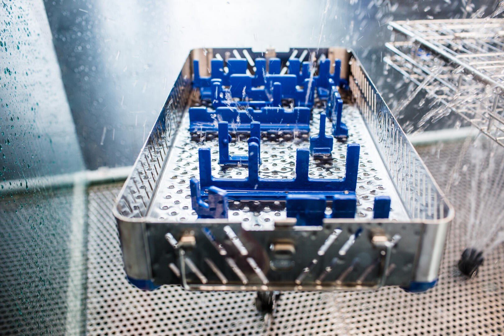 A metal tray with blue plastic supports is being cleaned with water jets inside a sterilization or washing machine, likely used for medical or laboratory instruments.