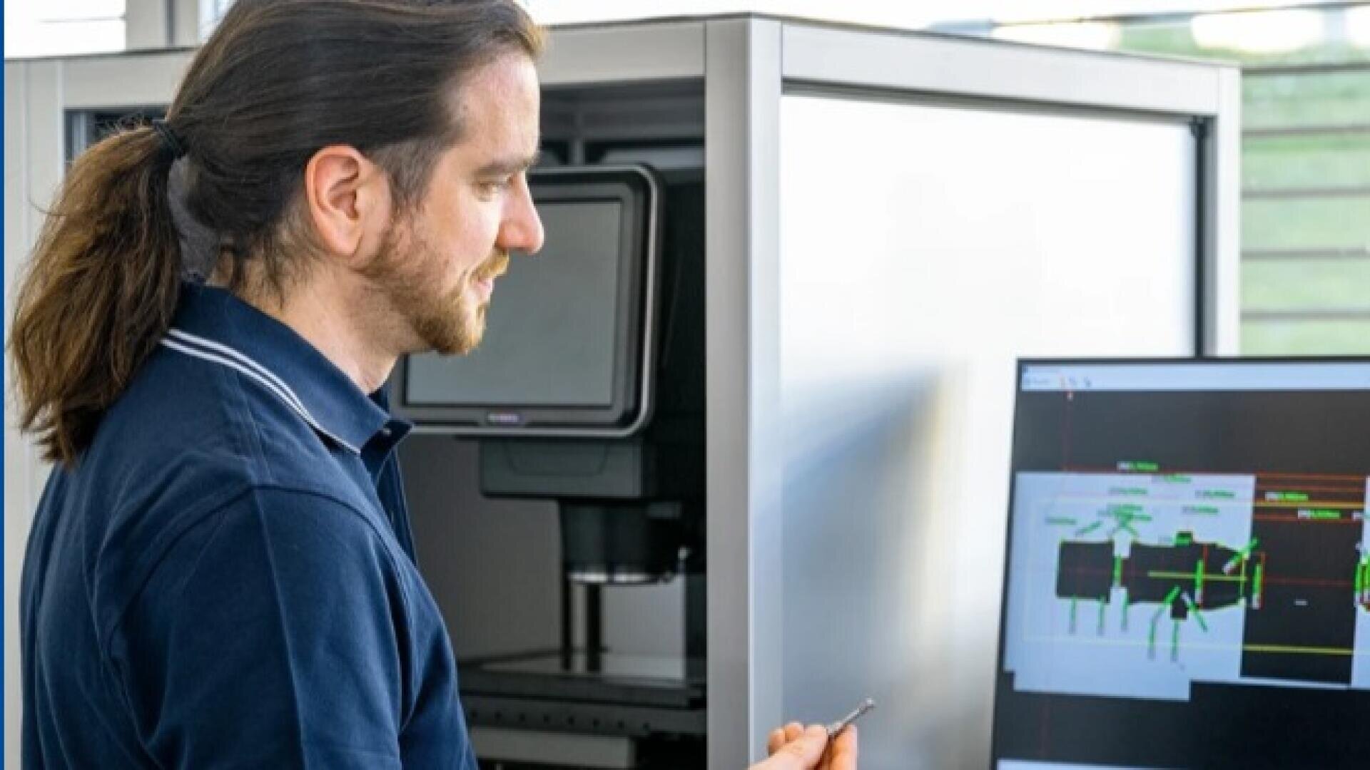 Ein Mann mit Pferdeschwanz und tätowiertem Arm hält einen kleinen Gegenstand in der Hand, während er auf einen Computerbildschirm mit technischen Diagrammen schaut. Er steht neben Laborgeräten in einem hellen, modernen Arbeitsraum.