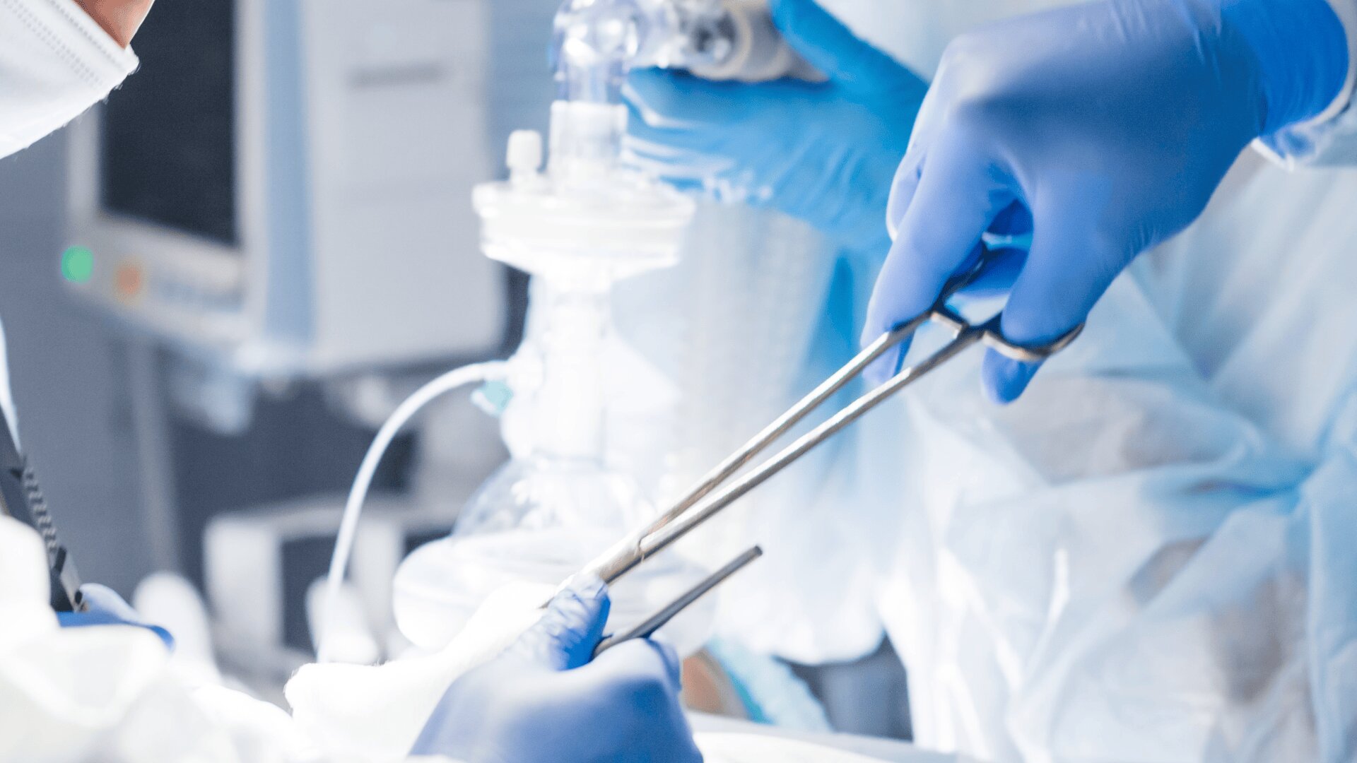 Close-up of surgeons wearing blue gloves and sterile gowns performing a medical procedure, holding surgical instruments in a brightly lit operating room with blurred medical equipment in the background.
