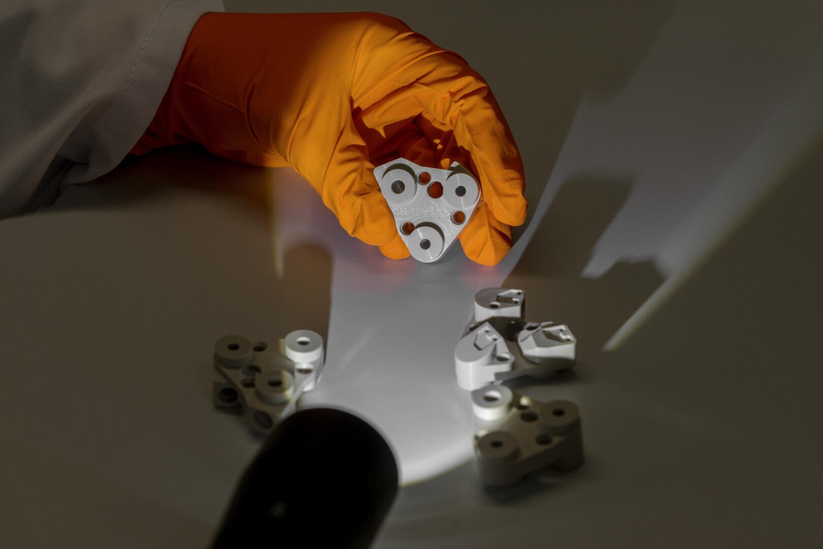 A person wearing an orange glove holds a small, white, irregularly shaped object with holes, while several similar objects lie on the table; a flashlight illuminates the scene and casts clear shadows.