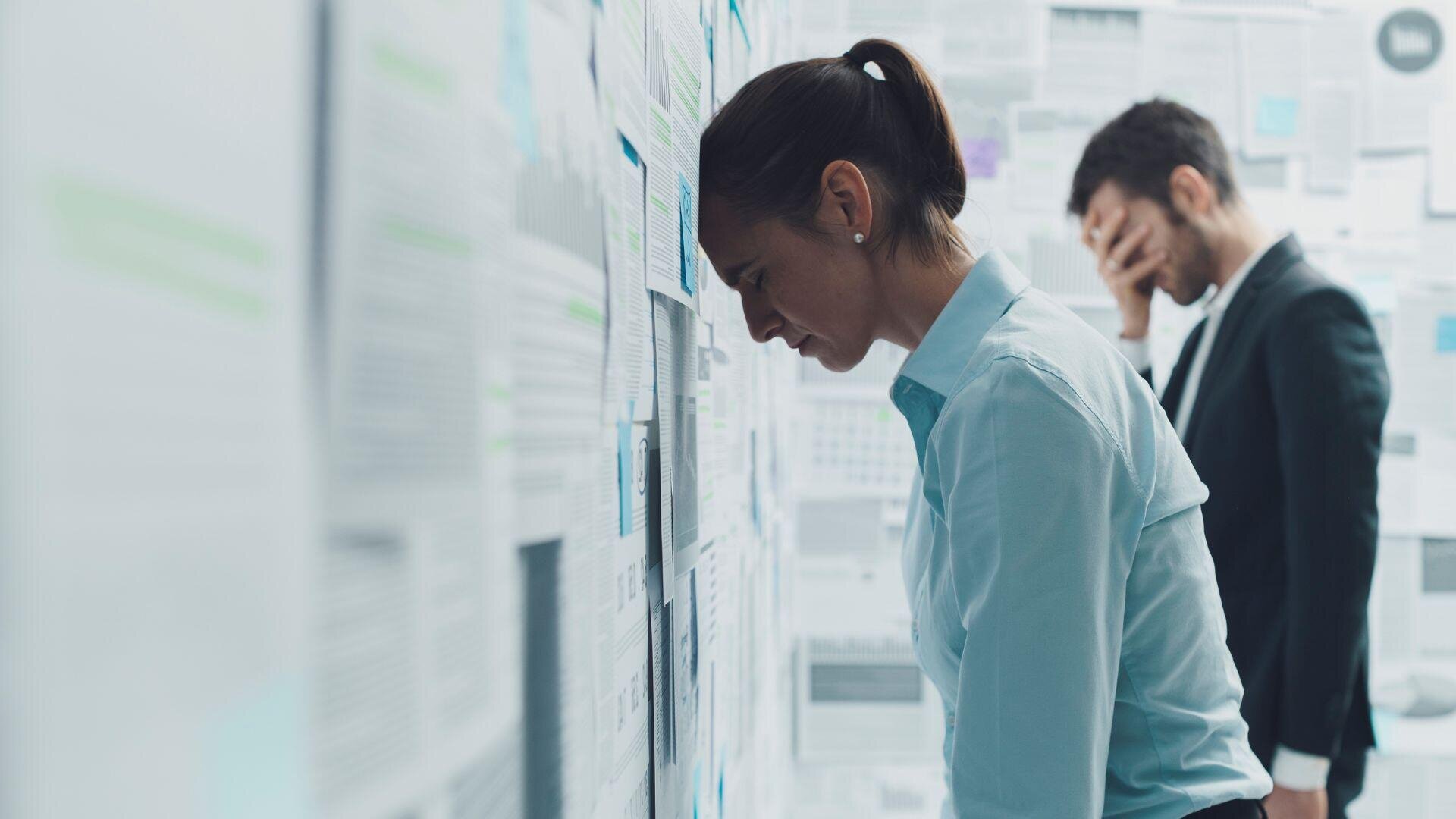 A woman in business attire leans her forehead in frustration against a wall covered with documents, while a man in the background covers his face with his hand and also appears stressed.