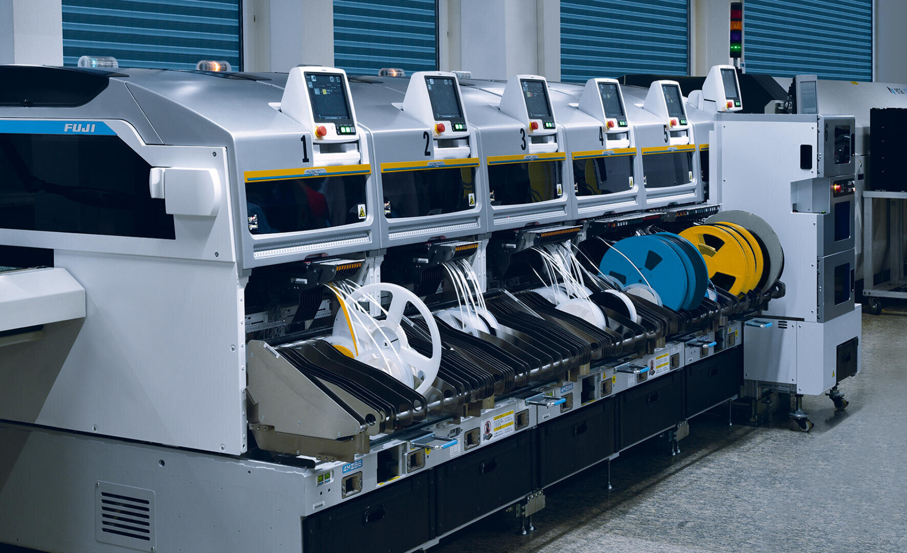 A row of automated Fuji machines on a factory floor, each with control screens and spools of different materials used to manufacture or assemble electronic components.