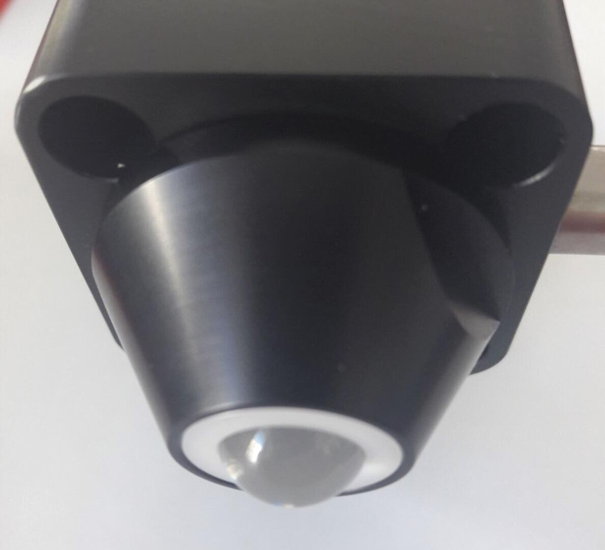 Nahaufnahme eines schwarzen zylindrischen Sensors oder einer Kamera mit einer markanten runden Linse oder Kuppel in der Mitte, die auf einem Metallarm montiert ist, vor einem schlichten weißen Hintergrund.