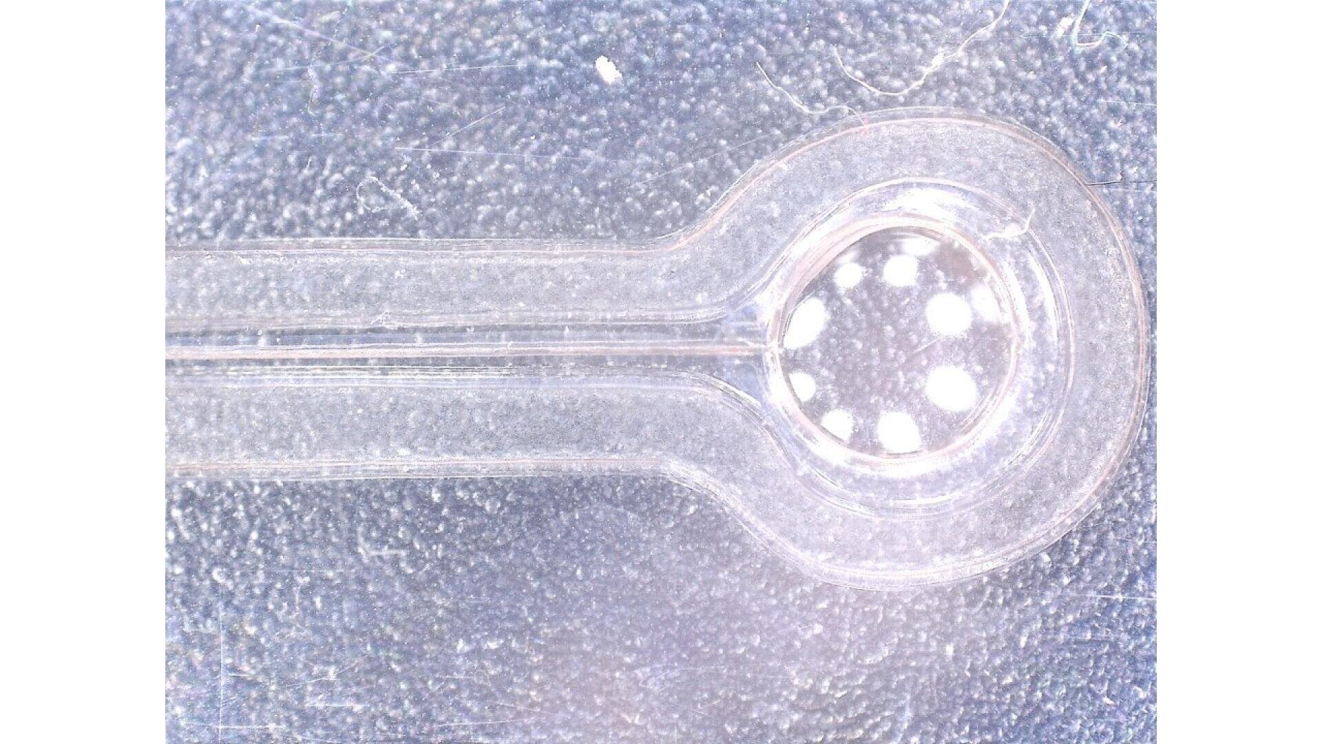 Close-up image of a transparent plastic object with a narrow handle leading to a rounded end containing six evenly spaced holes. The background is textured and softly lit.