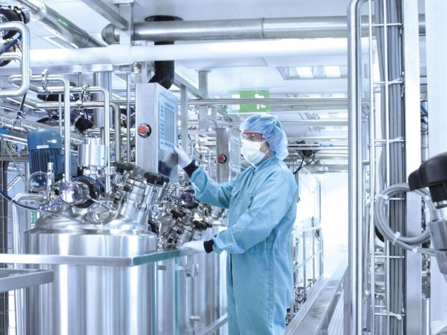 A person wearing protective clothing and a face mask operates machines in a sterile high-tech laboratory or pharmaceutical production plant, surrounded by stainless steel tanks and pipes.