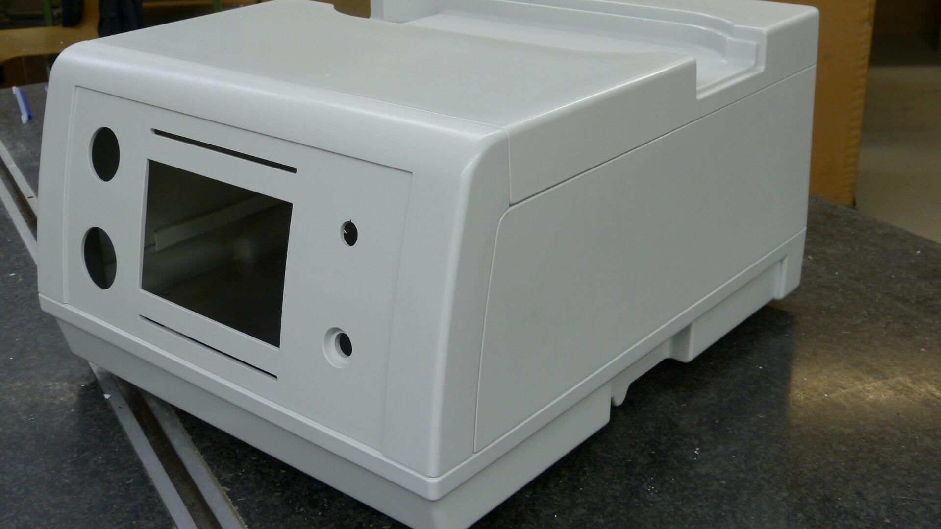 Ein weißes, rechteckiges Gehäuse für ein elektronisches Gerät mit einem nach vorne gerichteten Bildschirmausschnitt, runden und geschlitzten Öffnungen, das auf einer dunklen Oberfläche steht und für den Einbau von Komponenten vorbereitet ist.