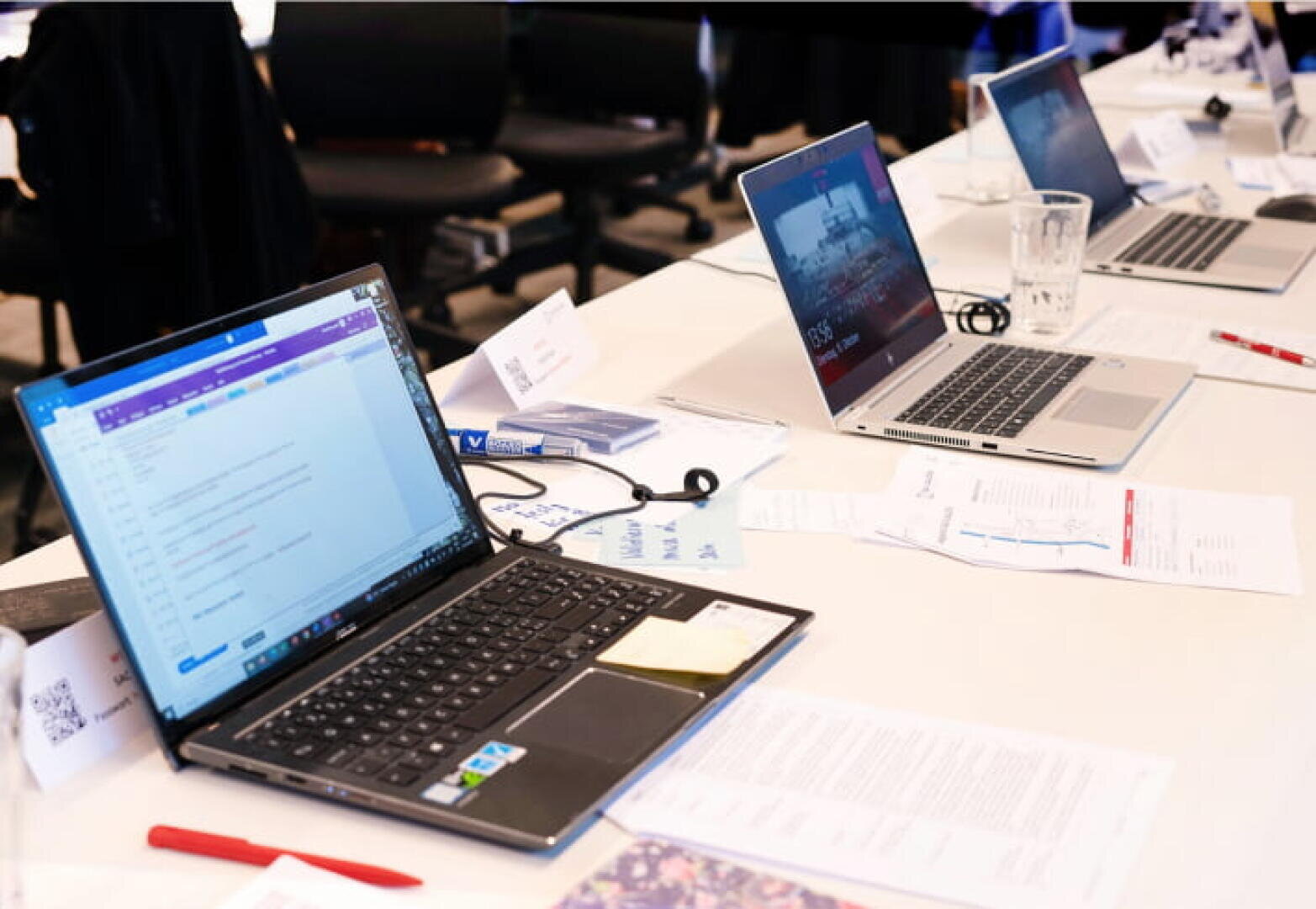Ein Konferenztisch mit offenen Laptops, Dokumenten, Wassergläsern, Stiften und Namenskarten. Die Laptops zeigen verschiedene Bildschirme an, und Papiere liegen verstreut herum, was auf eine Besprechung oder eine gemeinsame Arbeitssitzung hindeutet.