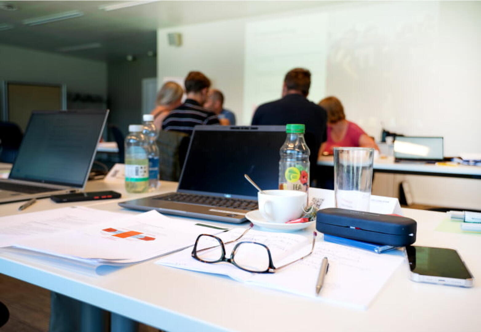 Eine Nahaufnahme eines Schreibtisches mit Papieren, einem Laptop, Gläsern, einer Tasse Kaffee und Wasserflaschen; im Hintergrund sitzen Menschen und sind unscharf, was auf eine laufende Sitzung oder einen Workshop hindeutet.