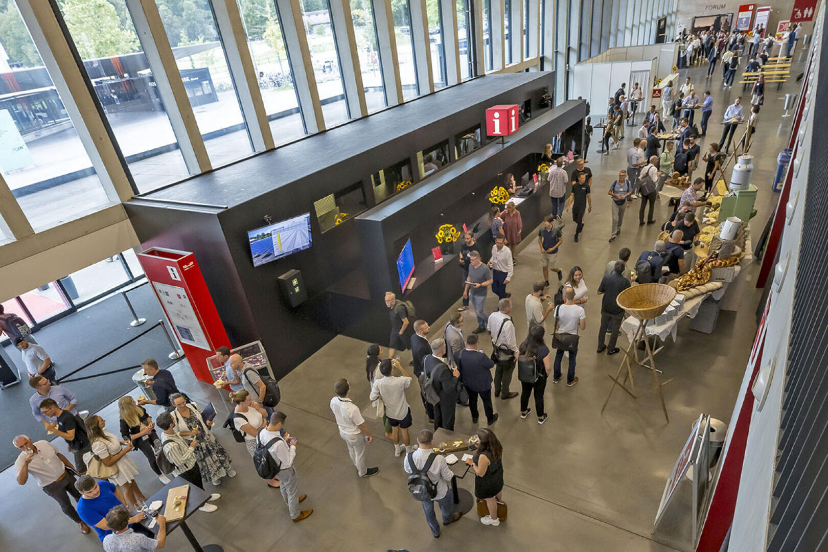 Eine große Gruppe von Menschen versammelt sich in einer geräumigen, modernen Lobby mit hohen Fenstern, mischt sich und genießt das Essen an einem Buffet, das wie eine Konferenz- oder Veranstaltungsanmeldung aussieht.