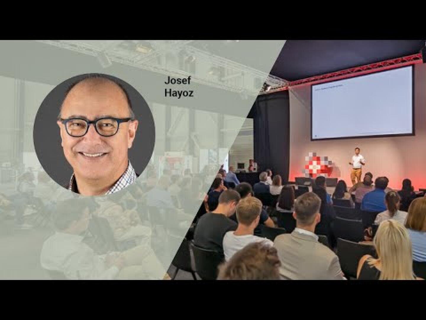 A photo from a conference with an audience facing a speaker on stage. On the left is a large overlay of a smiling man wearing glasses with the inscription Josef Hayoz. The event features a projected screen and red light accents.