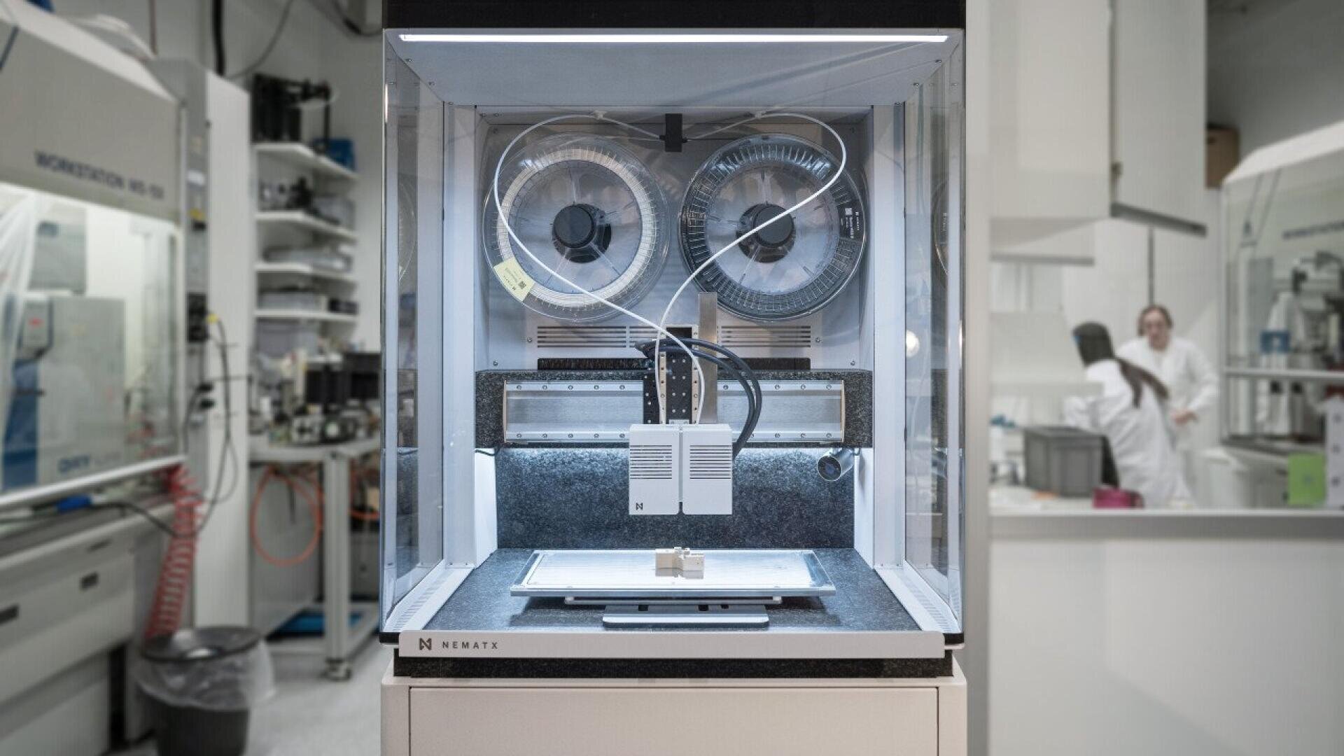 A 3D printer enclosed in a glass case operates in a modern laboratory, with spools of filament visible inside and lab equipment and people in white lab coats in the background.
