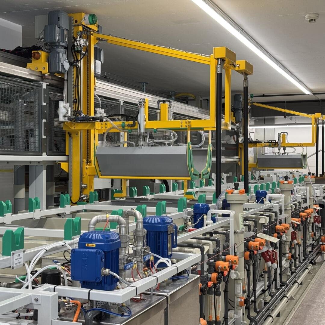 Automated industrial production line with mechanical arms, tanks, pipes, pumps, and control systems in a well-lit facility. The facility appears to be designed for chemical processing or manufacturing.