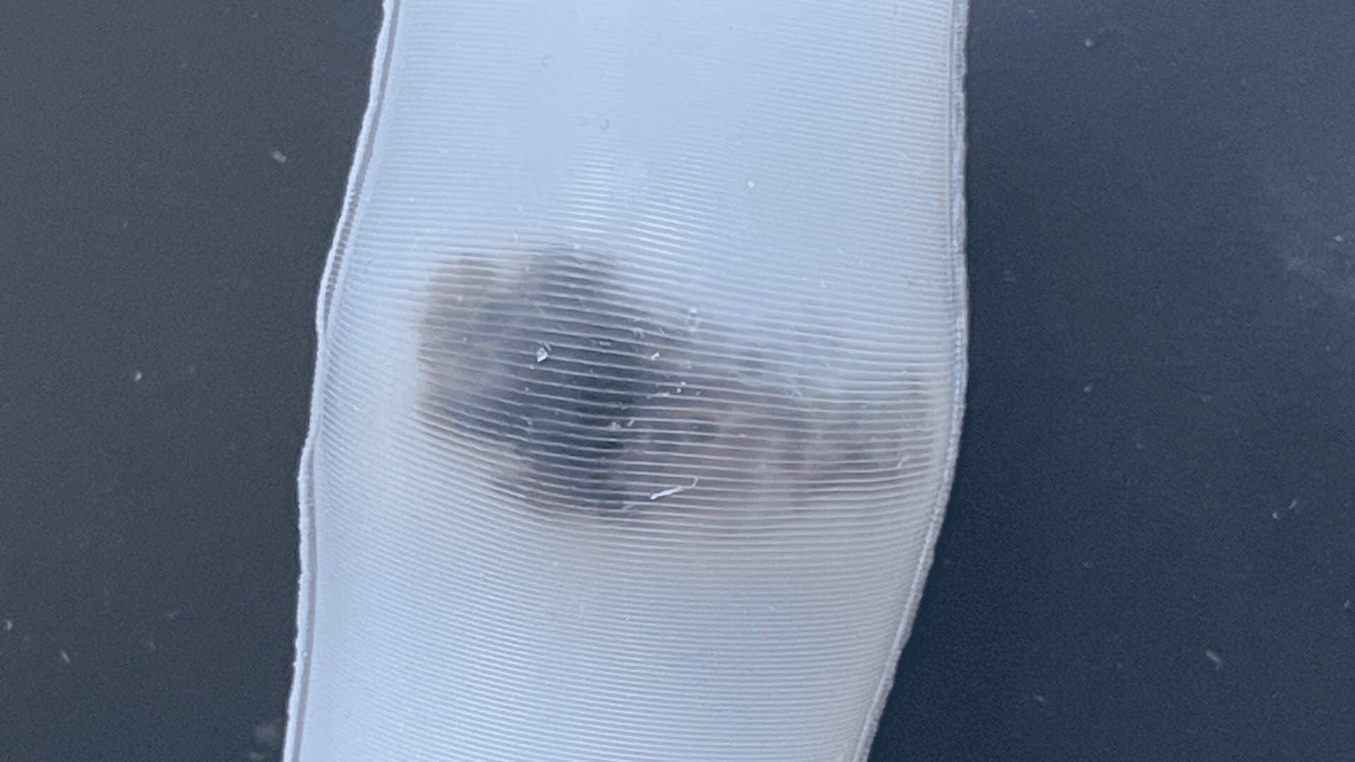 Nahaufnahme einer durchscheinenden, flexiblen Kunststoffoberfläche, unter der ein dunkler, unregelmäßiger Fleck zu sehen ist, der durch die Textur und die Krümmung des Materials leicht verzerrt ist.