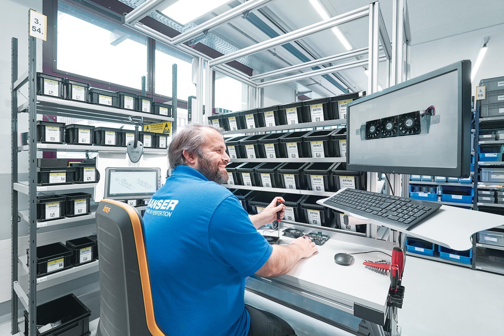A man in a blue shirt sits at a computer workstation surrounded by orderly shelves of labeled plastic bins in a bright, modern industrial setting.