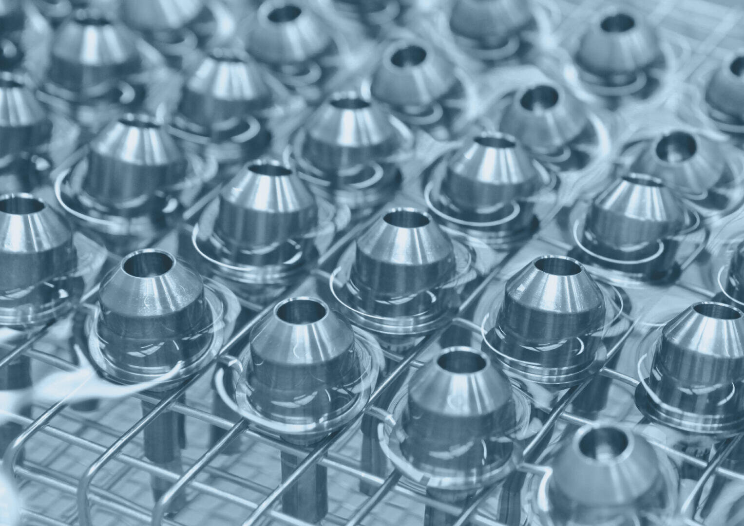 Nahaufnahme mehrerer glänzender Maschinenteile aus Metall, die in einem Gittermuster auf einem Drahtgestell angeordnet sind, wodurch ein sich wiederholendes und industrielles Aussehen entsteht.