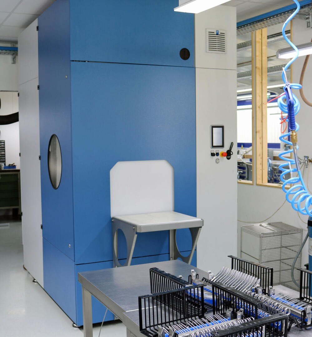Eine moderne Industriemaschine mit blau-weißem Äußeren steht in einer sauberen, hellen Fabrik. Im Vordergrund befinden sich Regale mit organisierten Metallwerkzeugen oder Komponenten. Der Bereich wirkt aufgeräumt und gut beleuchtet.