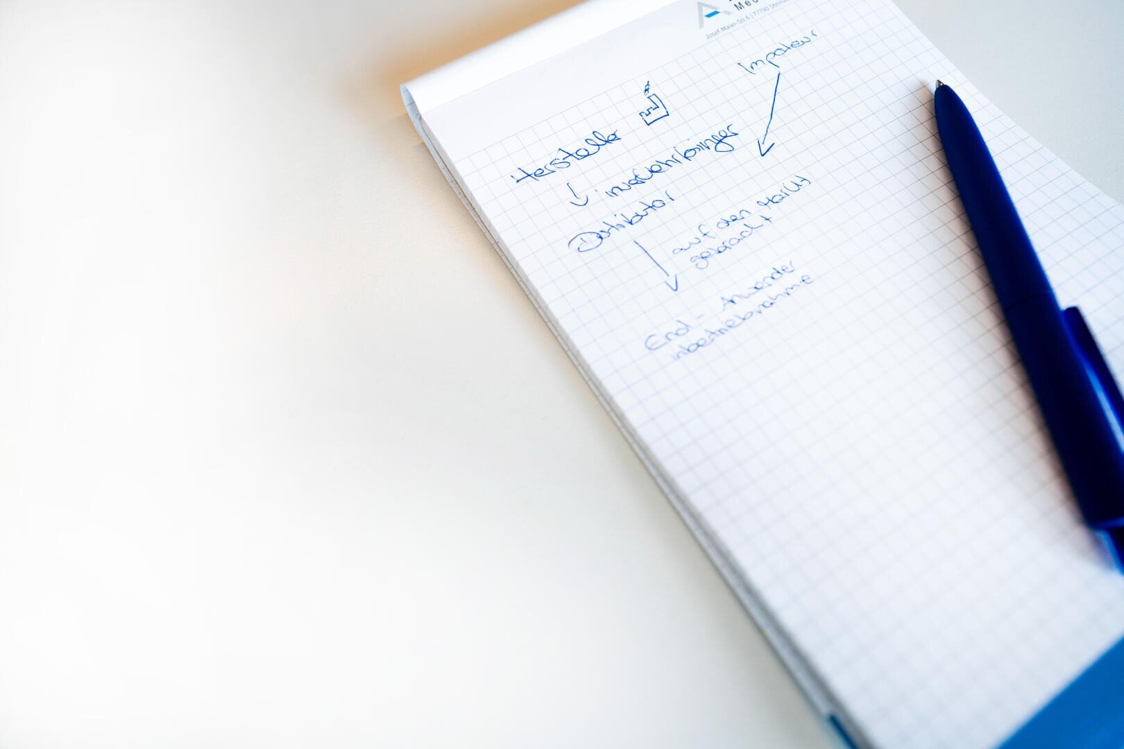 Ein blauer Stift ruht auf einem karierten Notizblock mit handschriftlichen blauen Notizen und Häkchen, der auf einem weißen Schreibtisch mit viel freiem Platz auf der linken Seite liegt.