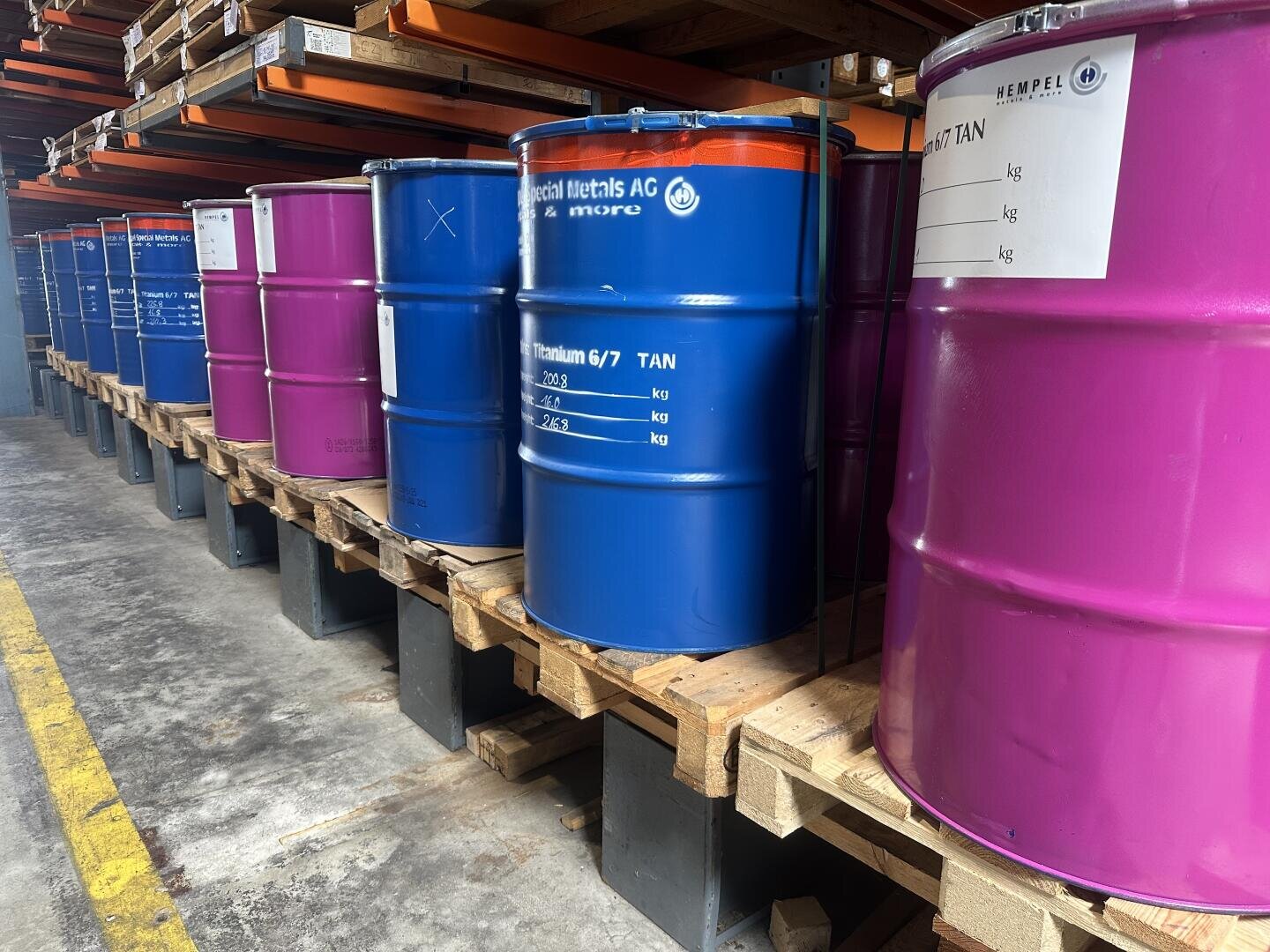 Rows of blue and purple industrial barrels are lined up on wooden pallets in a warehouse, with shelves above and a concrete floor below. Labels and markings can be seen on some of the barrels.