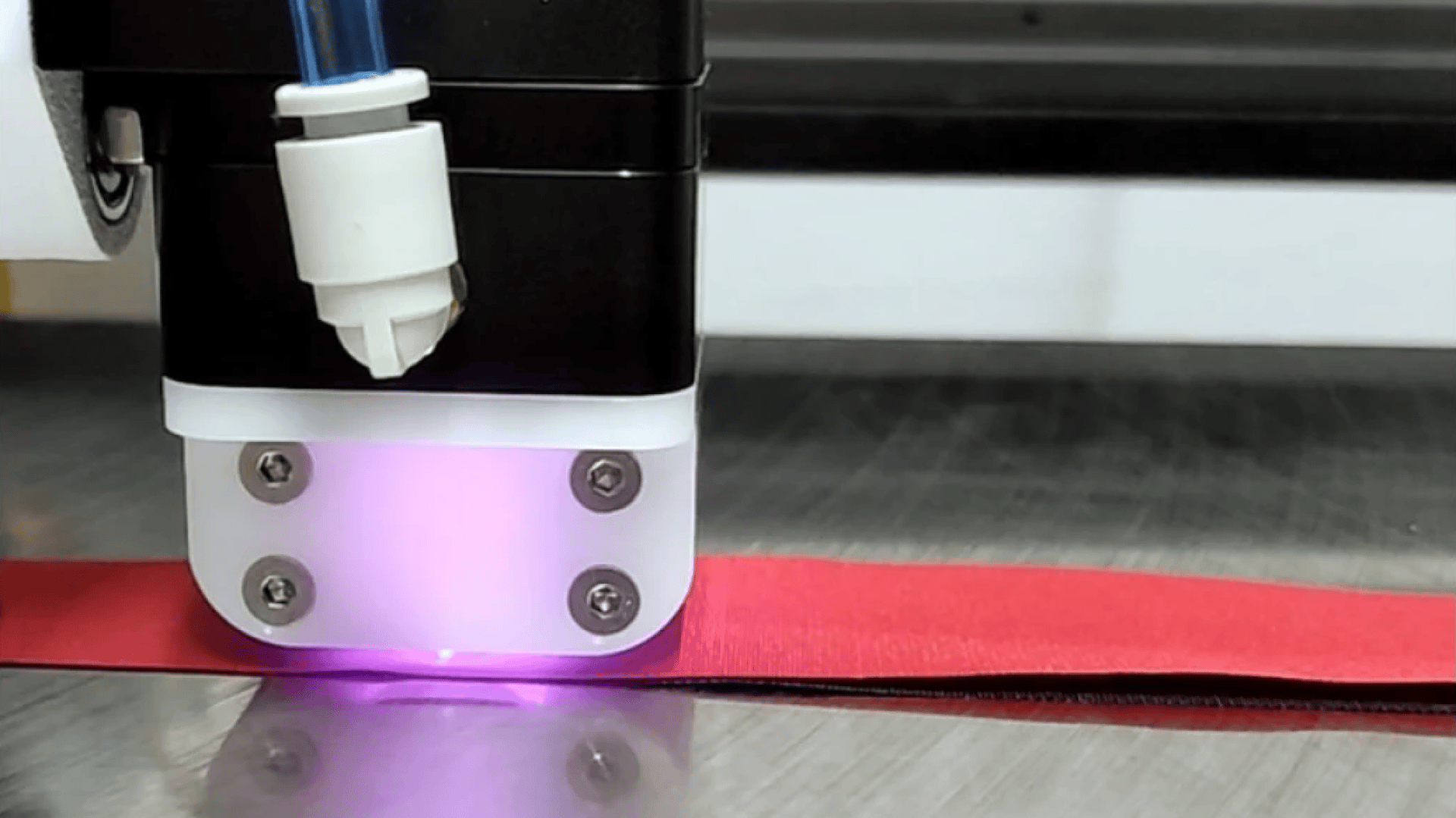 Nahaufnahme einer Laserschneidmaschine, die ein violettes Licht ausstrahlt, während sie einen roten Materialstreifen auf einer Metalloberfläche bearbeitet.