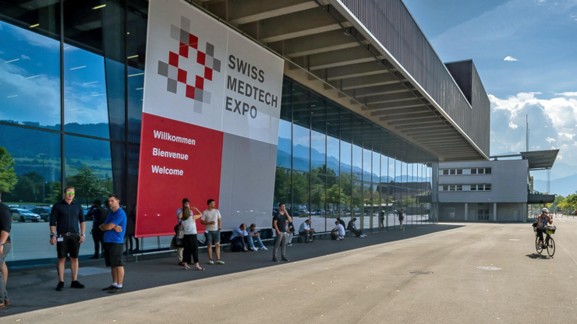 Menschen stehen und sitzen vor einem modernen Glasgebäude mit einem großen Schild mit der Aufschrift Swiss Medtech Expo und "Willkommen, Bienvenue, Welcome". Im Hintergrund sind Berge und blauer Himmel zu sehen.