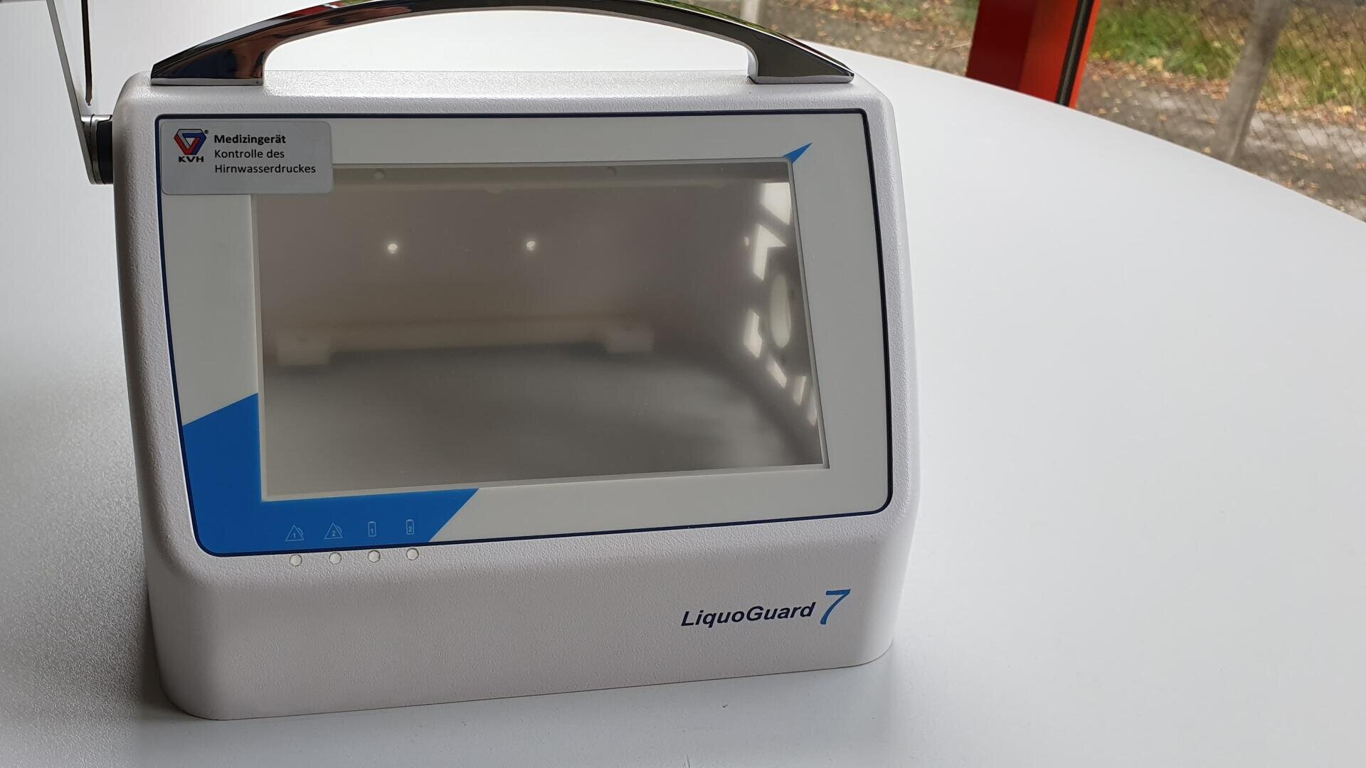 A white medical device labeled LiquoGuard 7 with a large screen, blue and gray accents and a handle lies on a round white table near a window. A sticker with German text is in the upper left corner.