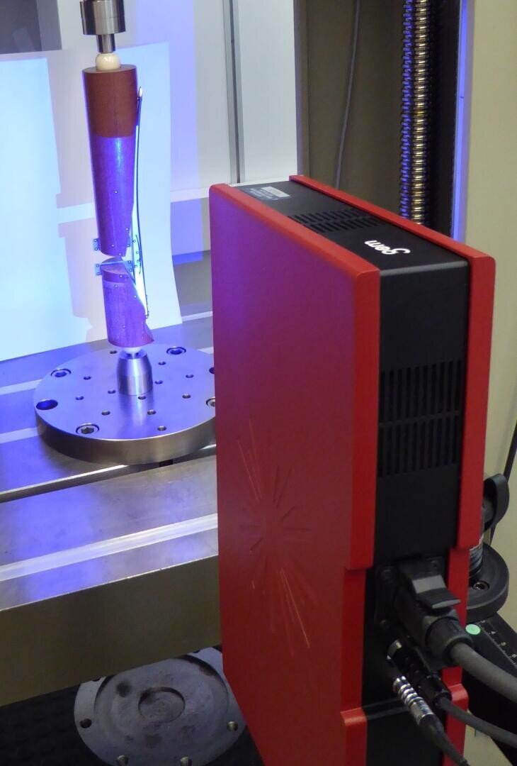 A red 3D scanner is positioned in front of a metallic object mounted vertically on a turntable for digital scanning, with wires connected to the scanner.
