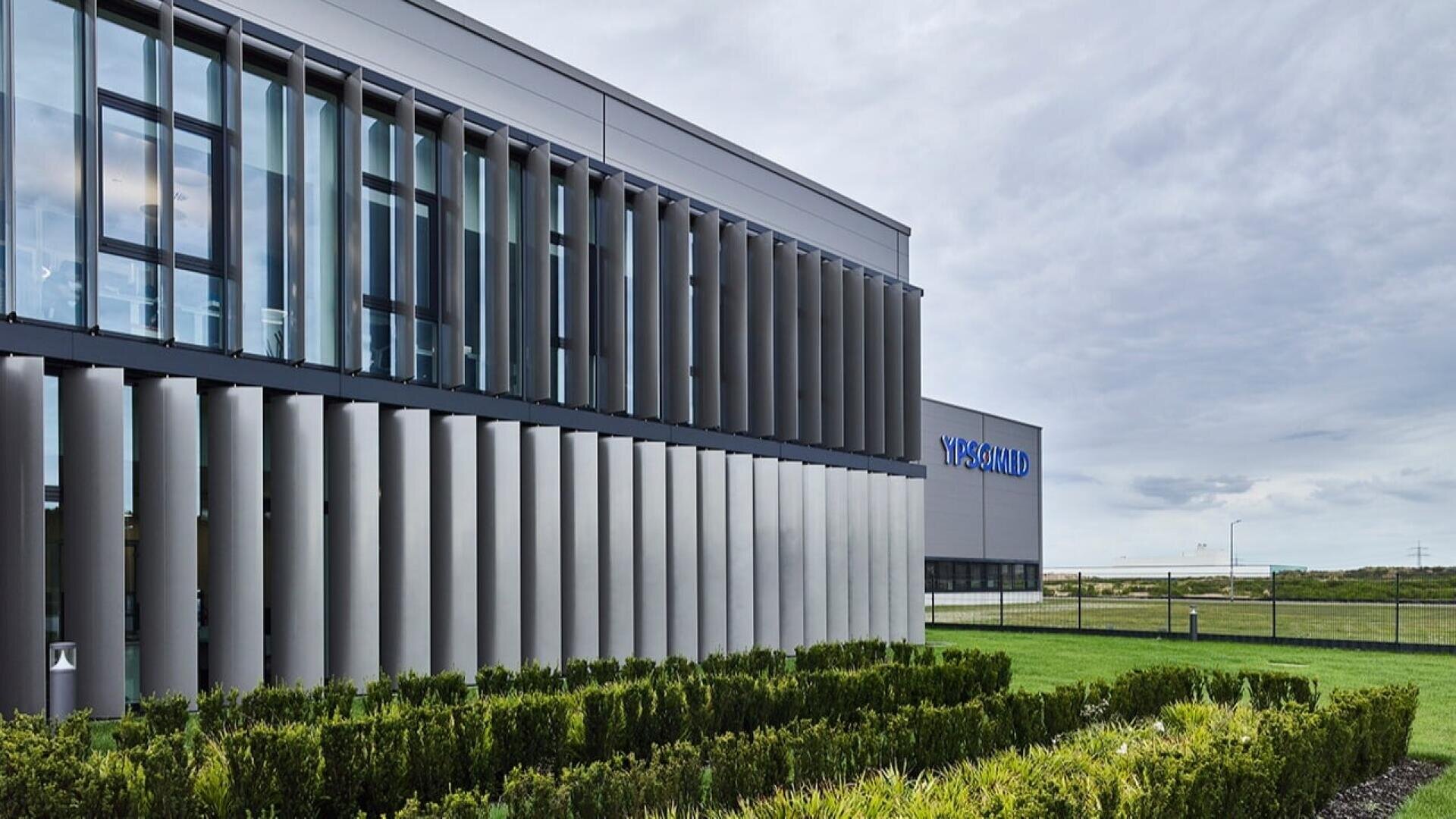 Modernes Industriegebäude mit vertikalen Metallpaneelen an den Fenstern, ordentlich getrimmte Büsche im Vordergrund und ein bewölkter Himmel. Das Wort VIRSAMO ist auf dem Gebäude im Hintergrund zu sehen.