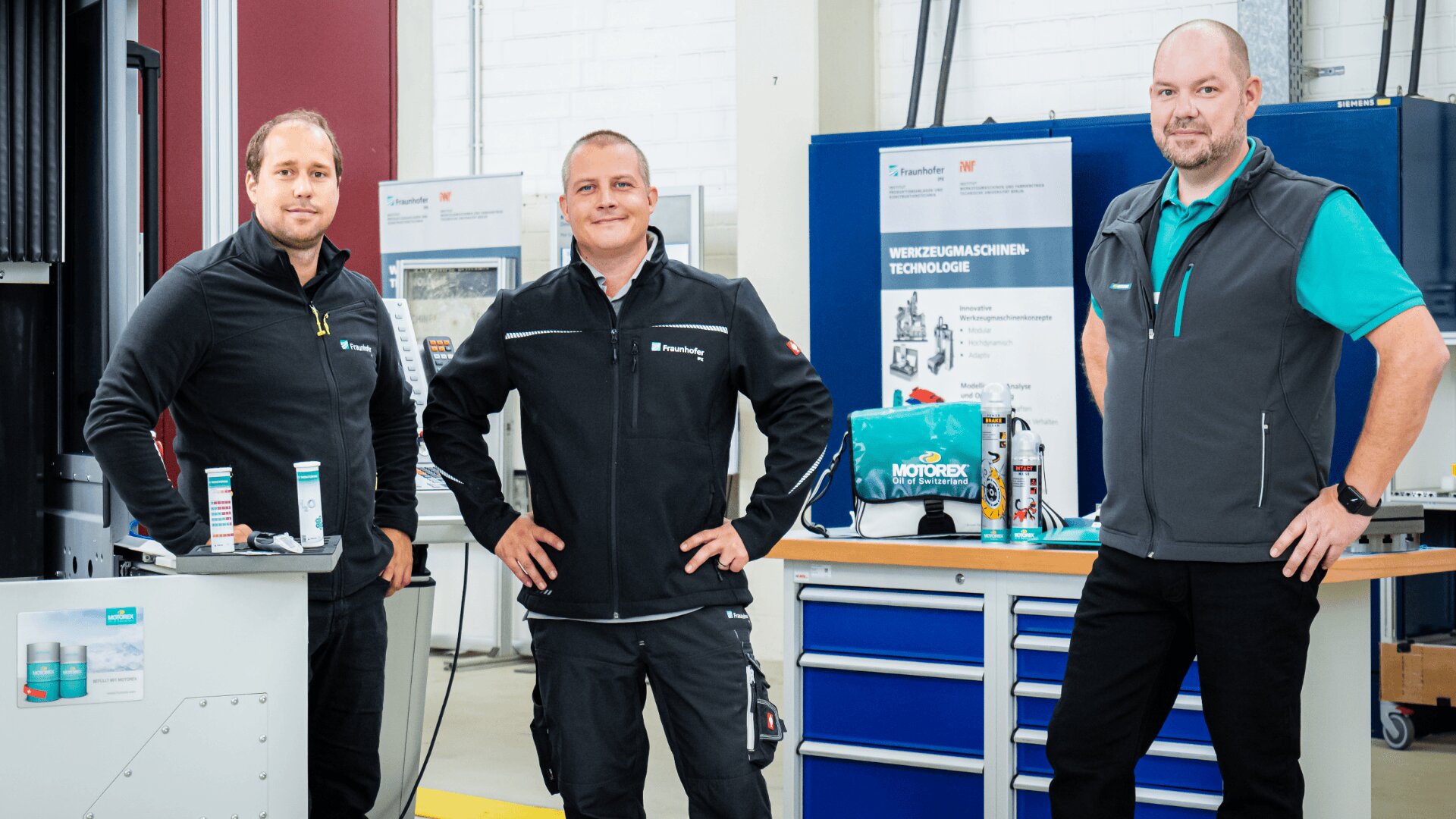 Drei Männer in schwarzen Jacken stehen in einer Industriehalle, posieren und lächeln in die Kamera. Im Hintergrund sind Werkbänke, Werkzeuge und Informationsplakate zu sehen.