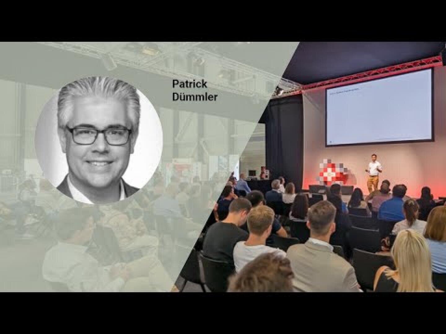 Ein Redner spricht auf der Bühne vor einem Publikum in einem Konferenzraum. Auf der linken Seite ist ein schwarz-weißes Kopfbild eines Mannes mit der Aufschrift "Patrick Dümmler" eingeblendet. Das Publikum hört aufmerksam zu.