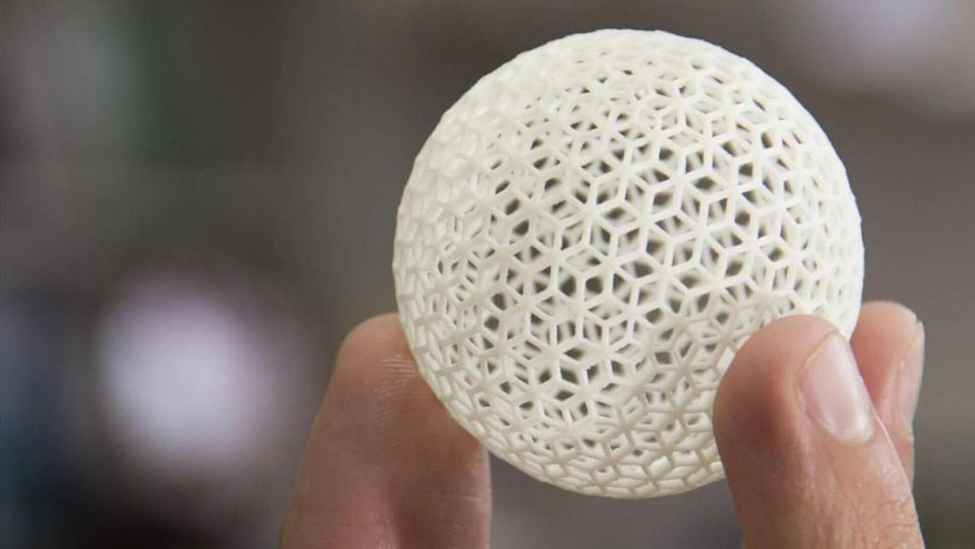 Eine Hand hält eine weiße 3D-gedruckte Kugel mit einem komplizierten geometrischen Gittermuster, das feine Details und Texturen vor einem unscharfen Hintergrund zeigt.