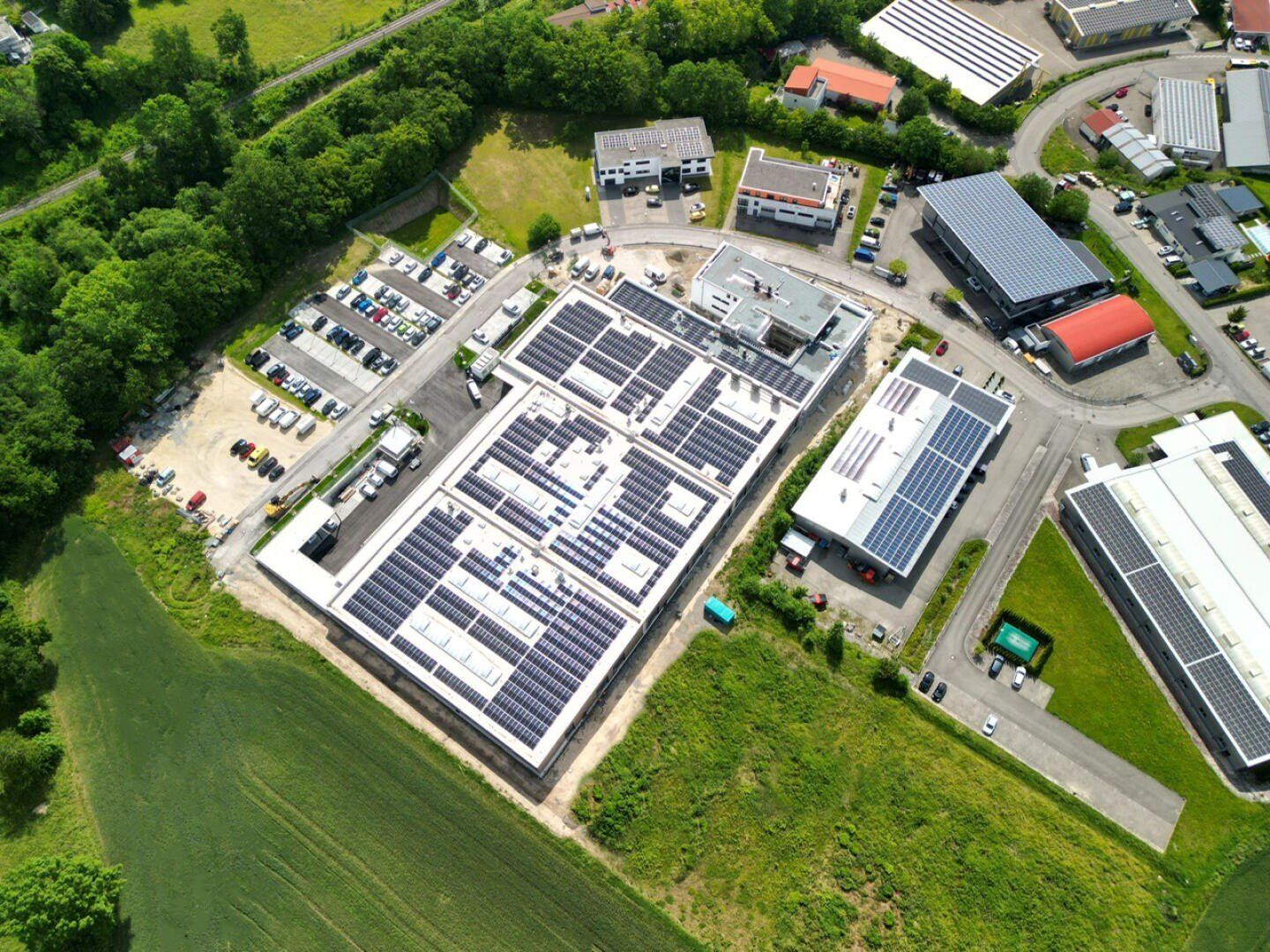 Aerial view of an industrial complex with several buildings with solar panels on the roofs, surrounded by parking lots, green fields and trees.