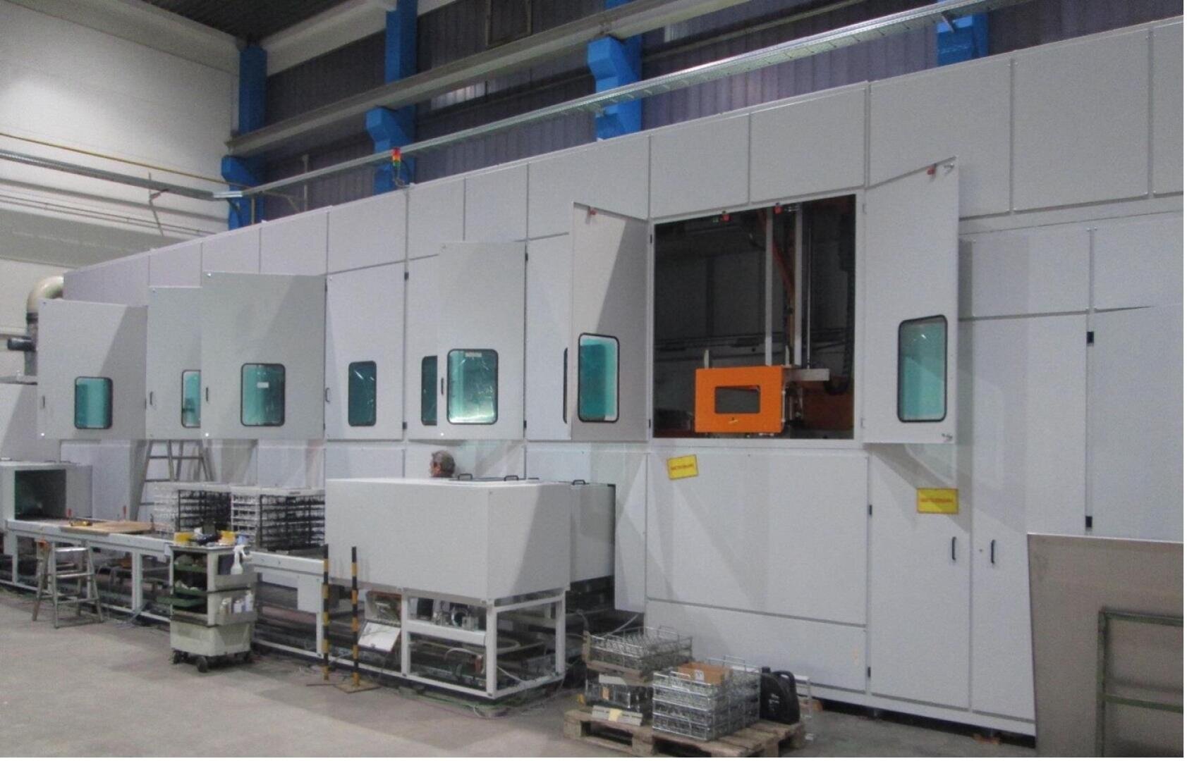 Eine große Industriemaschine mit mehreren weißen Platten und grünen Fenstern steht in einer Fabrik. Einige Paneele sind geöffnet und geben den Blick auf die Geräte im Inneren frei. Metallgestelle, Wagen und Tabletts sind um die Maschine herum angeordnet.