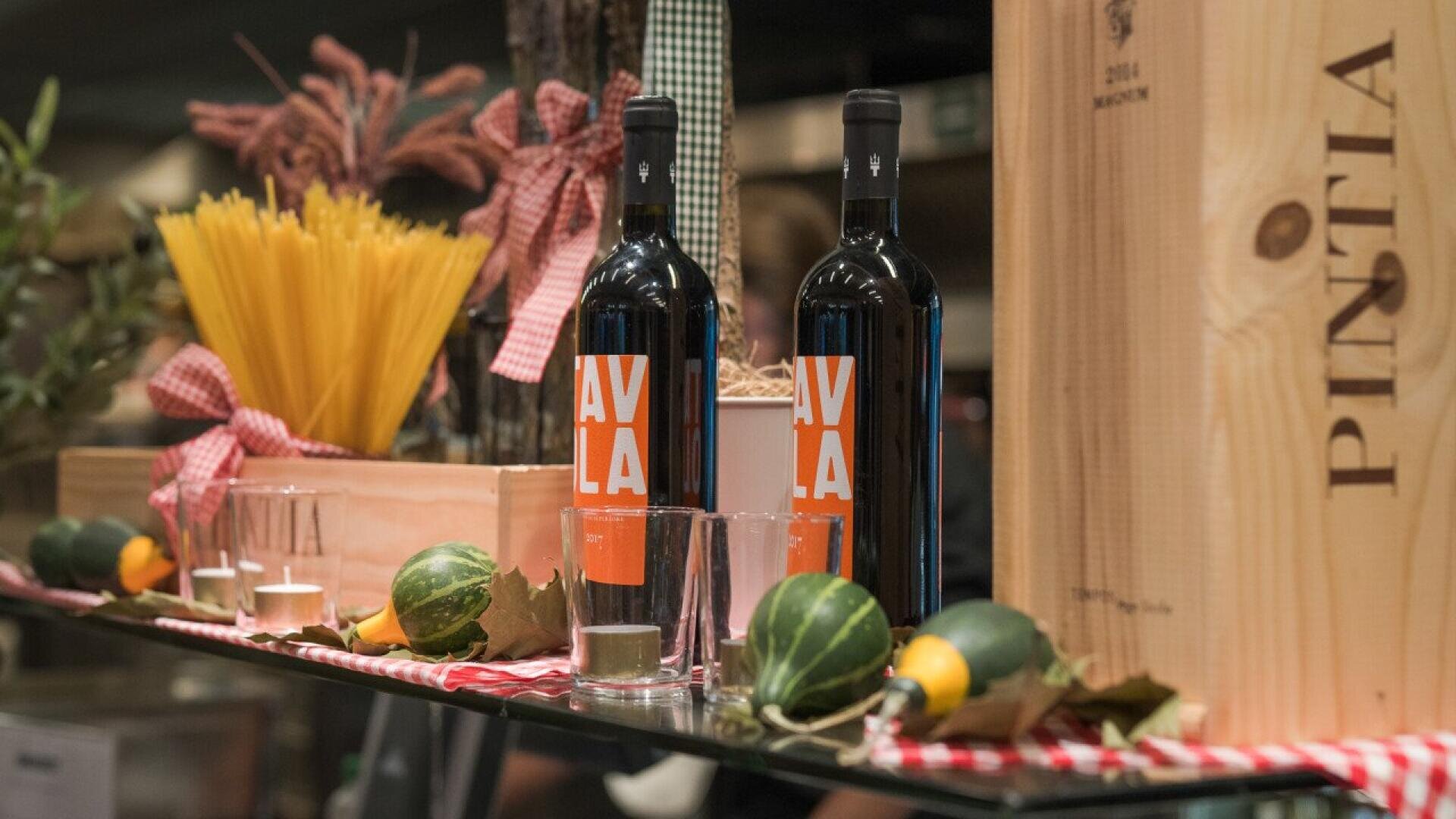 Two bottles of red wine sit on a glass shelf alongside glasses, decorative squash, a crate of uncooked spaghetti, candles, and a large wooden wine box labeled Pintia. A red gingham cloth accents the display.