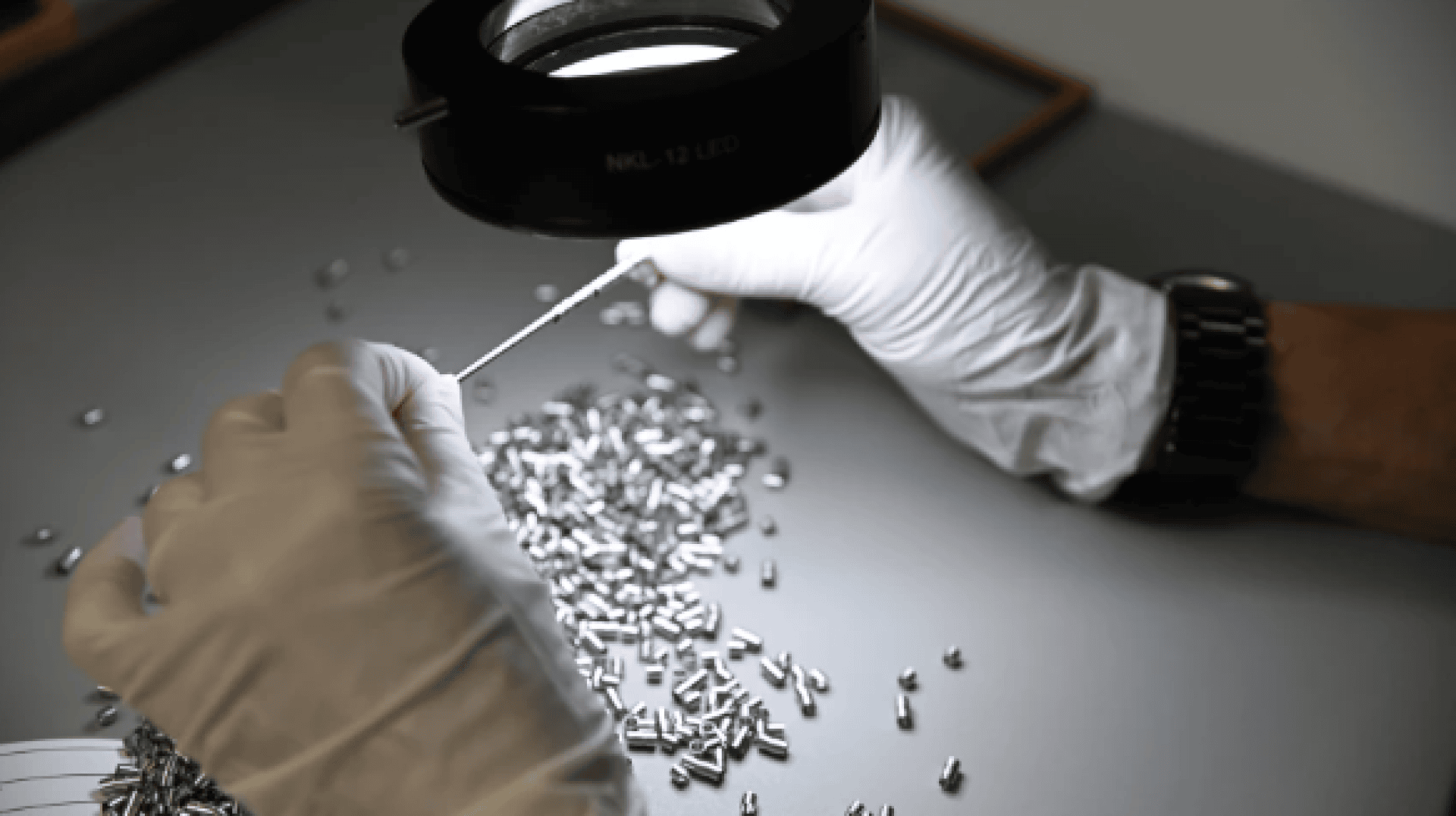 Eine Person mit weißen Handschuhen untersucht unter einer Vergrößerungslampe ein kleines Metallteil, auf dem viele ähnliche Metallteile auf einer grauen Fläche verstreut sind.