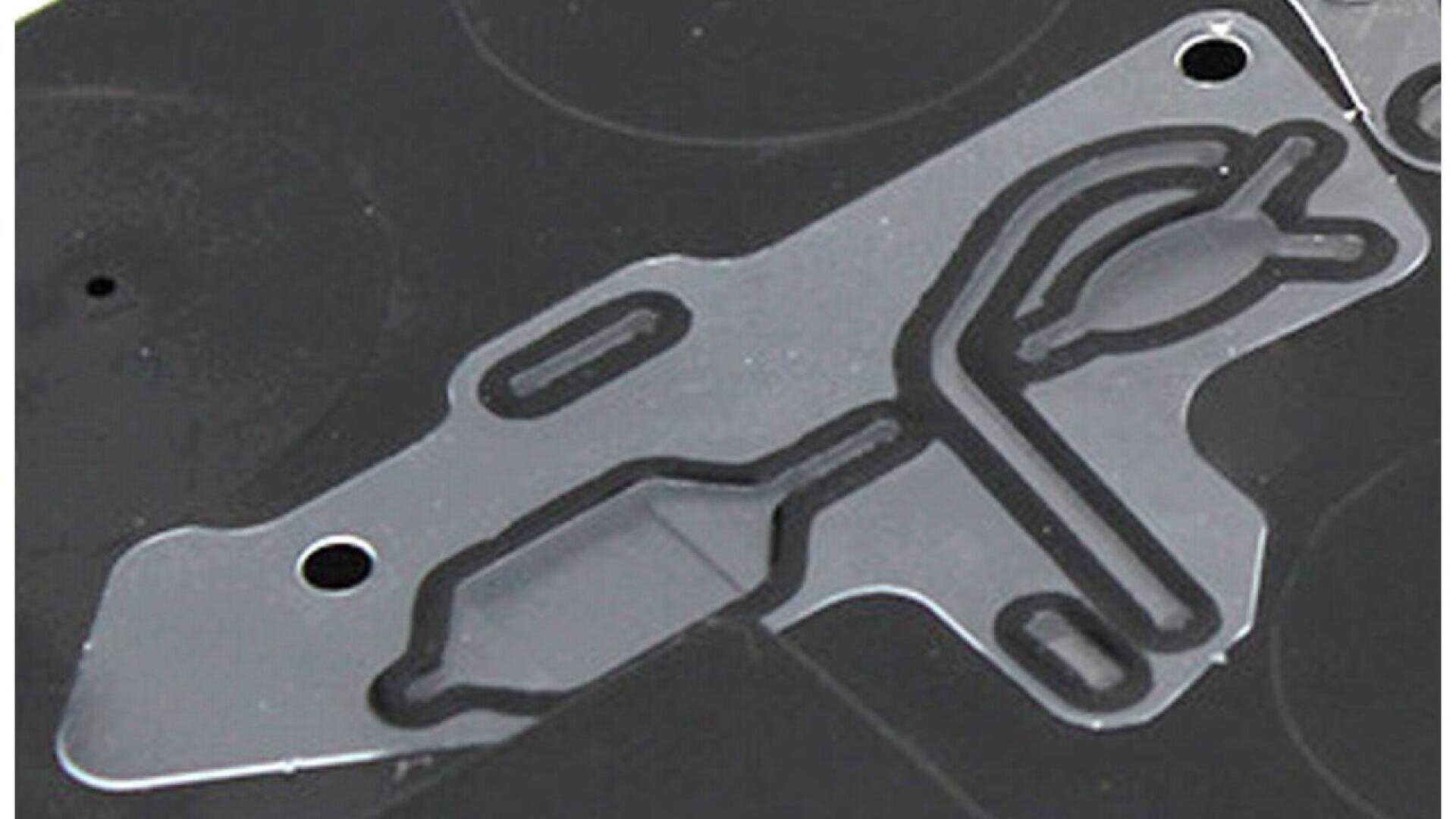 Close-up of a black and transparent plastic automotive gasket with intricate cutouts and grooves, showing two bolt holes and channel patterns for fluid or air passage.