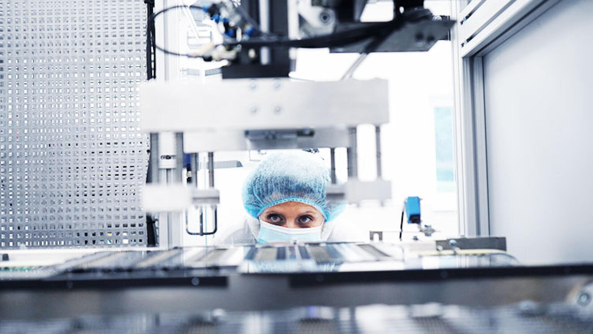 Ein Wissenschaftler mit Haarnetz, Gesichtsmaske und Laborkittel beobachtet ein großes Laborgerät in einer sauberen, hochtechnologischen Laborumgebung.
