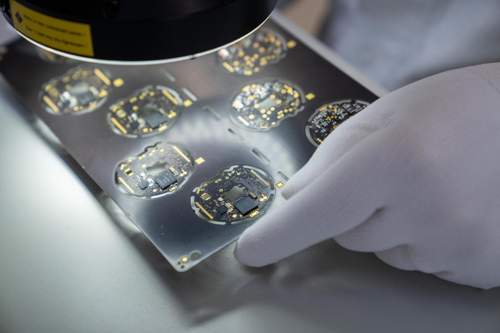 Eine behandschuhte Hand zeigt auf eine Metallschale, auf der sich mehrere runde elektronische Leiterplatten befinden, die mit einer Lupe untersucht werden.