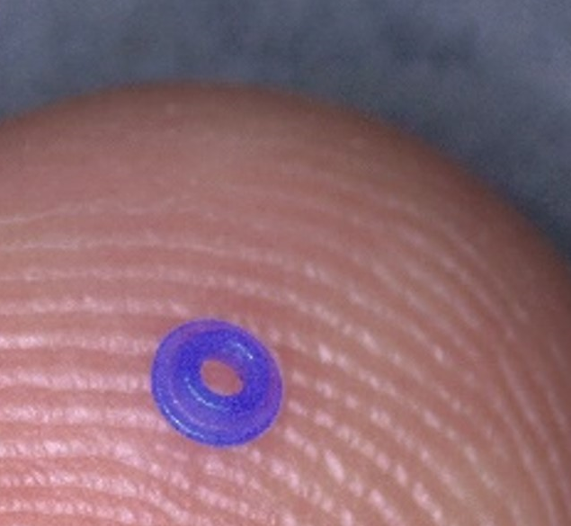 A tiny purple washer sits on the tip of a human finger, emphasizing its small size against the ridges of the fingerprint.