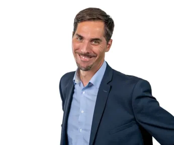 Ein Mann mit kurzen braunen Haaren und einem Ziegenbart lächelt, während er eine blaue Anzugsjacke und ein hellblaues Hemd trägt und vor einem schlichten weißen Hintergrund steht.