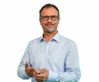 Ein Mann mittleren Alters mit Brille, kurzem dunklem Haar und gestutztem Bart lächelt freundlich. Er trägt ein hellblaues Hemd und steht vor einem schlichten weißen Hintergrund.