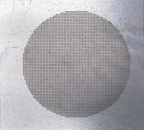 A round pattern of small holes arranged in a grid, forming a large circle on a metallic, brushed silver surface. The circle is centered and contrasts with the surrounding flat metal.