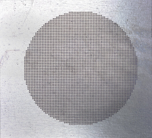 Ein großer Kreis aus kleinen Löchern ist auf einer metallischen, strukturierten Oberfläche zentriert. Der Hintergrund sieht aus wie gebürstetes Metall mit leichten Kratzern und einer reflektierenden, silbrigen Oberfläche.
