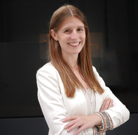 Eine Frau mit langen glatten hellbraunen Haaren lächelt mit verschränkten Armen. Sie trägt einen weißen Blazer, ein beiges Oberteil und Armbänder und steht vor einem dunklen, verschwommenen Hintergrund.
