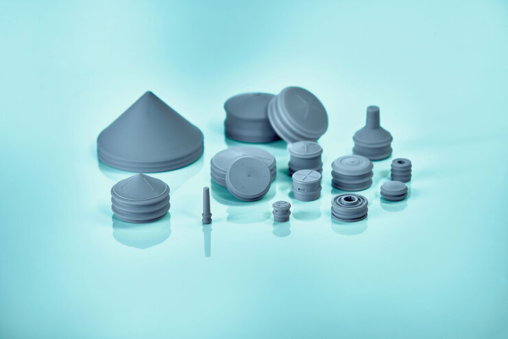 A collection of gray rubber stoppers, caps, and seals of various shapes and sizes is arranged on a reflective, light blue surface.