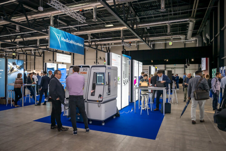Die Besucher nehmen an einer Messe in einer großen Halle teil und interagieren an verschiedenen Ständen mit Ausstellungsstücken, Bannern und Geräten. Die Atmosphäre ist professionell und konzentriert sich auf technologie- oder branchenbezogene Exponate.