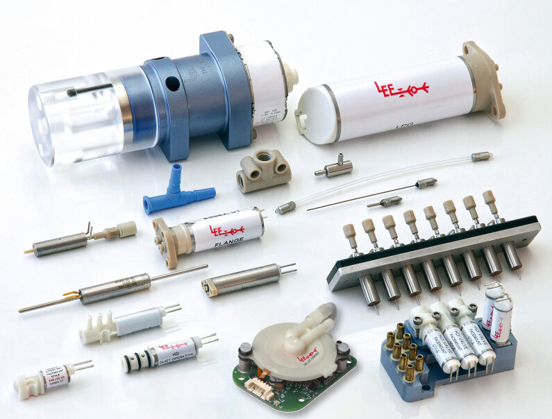 Verschiedene kleine industrielle und elektronische Bauteile, darunter Ventile, Röhren, Steckverbinder und Schaltkreisteile, angeordnet auf einer weißen Fläche. Auf mehreren Teilen sind Etiketten mit LEE- und technischen Kennzeichnungen zu sehen.