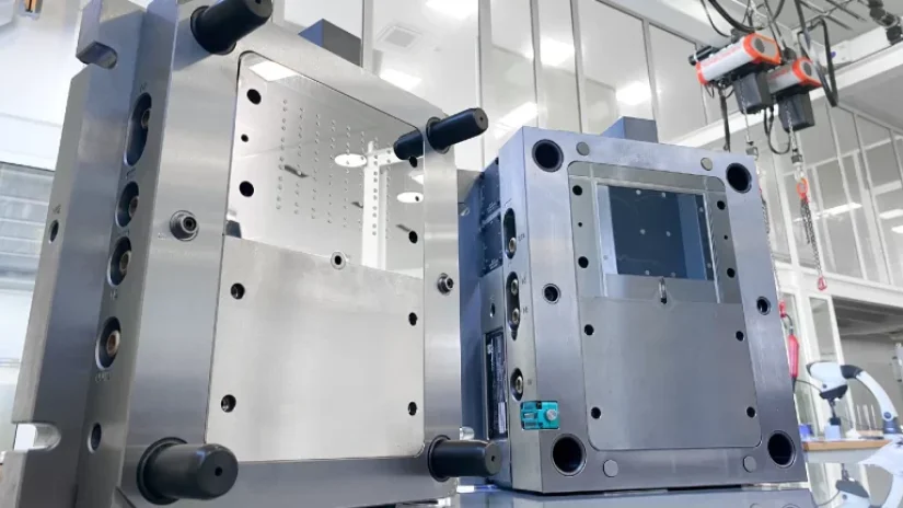 Two large metal injection molds are open, displaying smooth inner surfaces and various holes, set in a clean, well-lit industrial environment with machinery visible in the background.