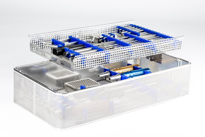 A metal mesh sterilization tray with two layers holds assorted surgical instruments, organized in blue silicone brackets, against a white background.