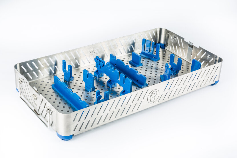 A stainless steel sterilization tray with perforated sides and bottom, featuring blue plastic brackets arranged inside to securely hold medical instruments. The tray is empty and placed on a white background.
